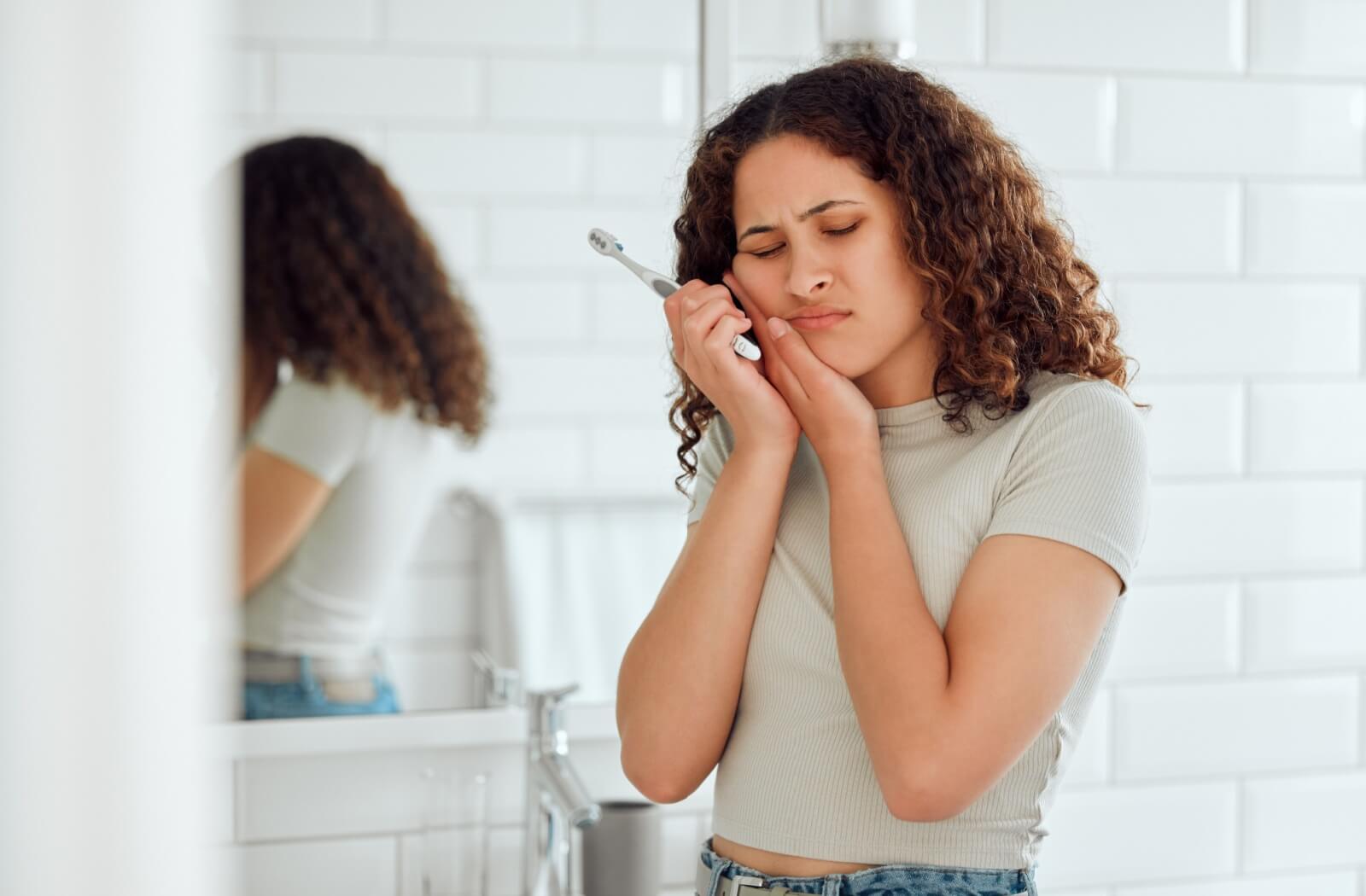 A woman holding her cheek in pain, indicating tooth discomfort while standing in a bathroom.